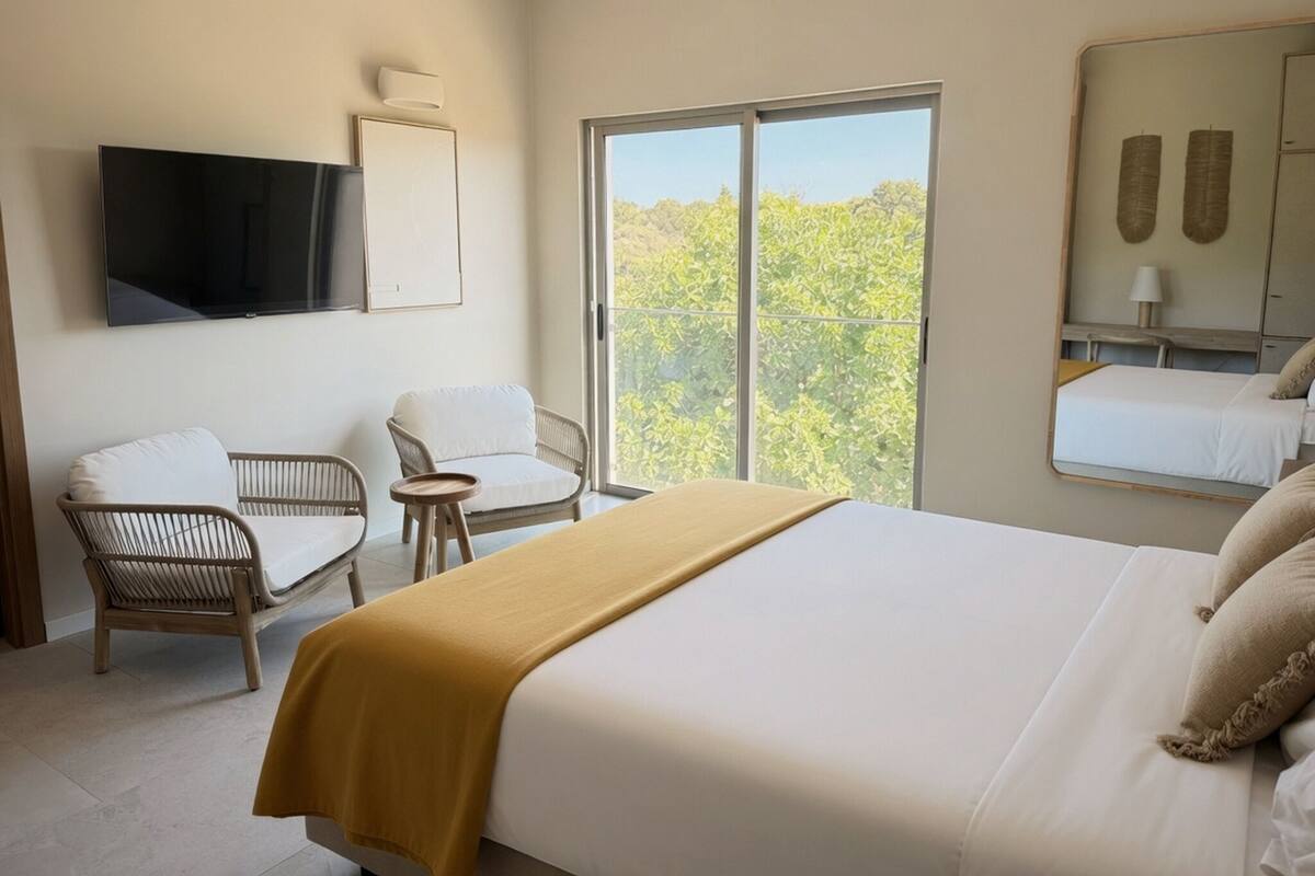 Habitación de hotel con cama, dos sillas, una mesita, televisor y un ventanal con vistas.