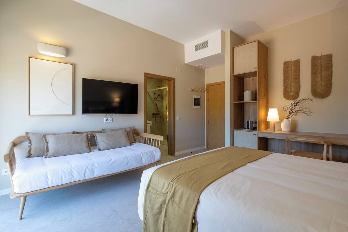 Una habitación de hotel con cama, sofá, televisor, escritorio con lámpara y estante con objetos decorativos.