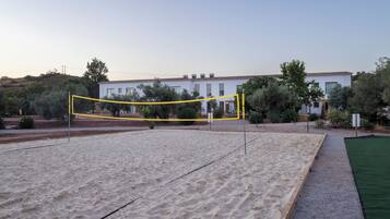 Una cancha de voleibol de arena con red, rodeada por un área ajardinada con árboles y un edificio al fondo.