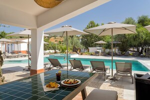 Zona junto a la piscina con sillones de descanso, sombrillas y una mesa con comida.