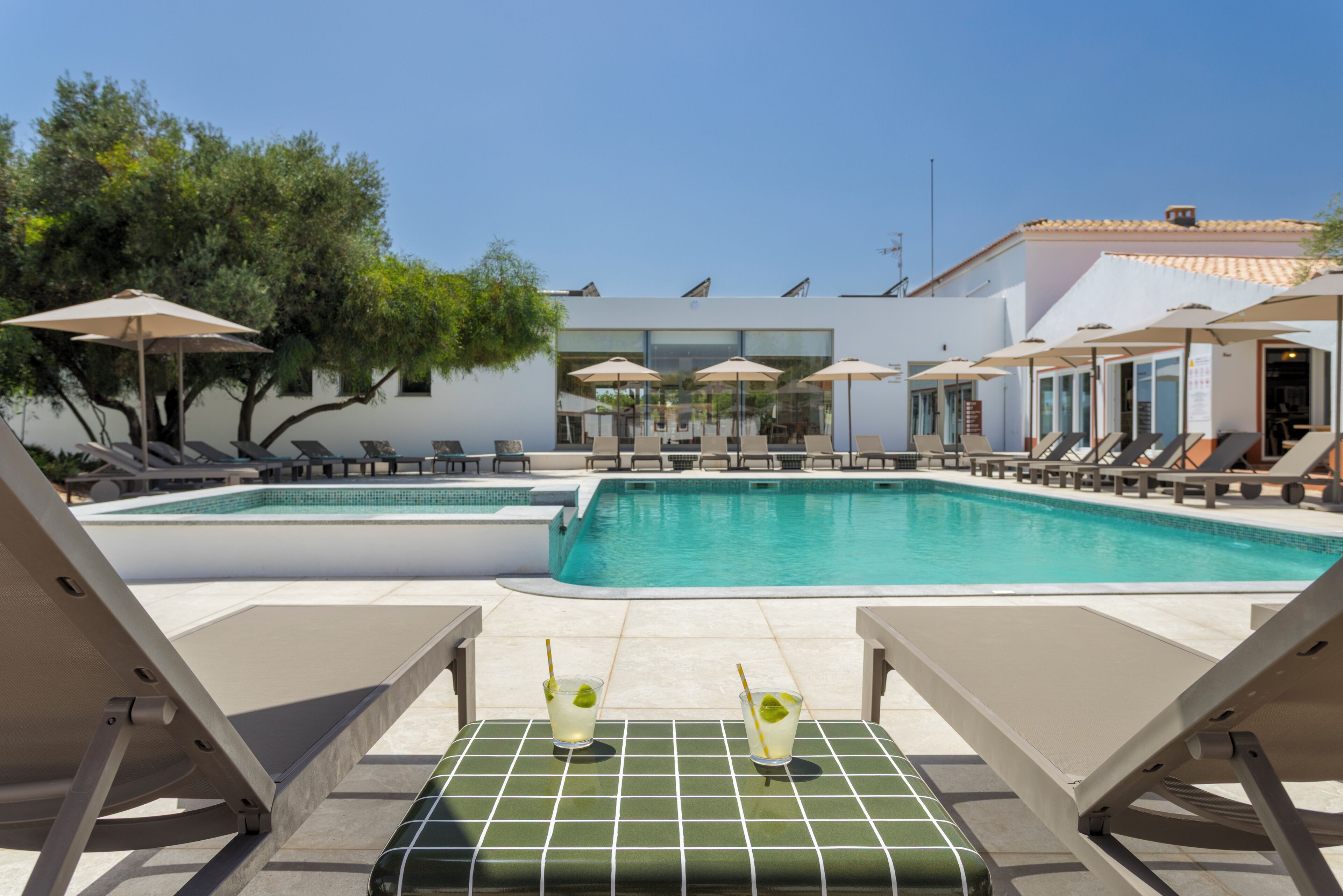 Ein Poolbereich mit Liegestühlen und Sonnenschirmen, ein Tisch mit Getränken und ein modernes Gebäude im Hintergrund.