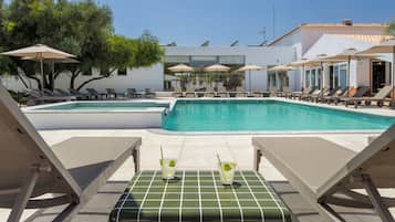 Ein Poolbereich mit Liegestühlen und Sonnenschirmen, ein Tisch mit Getränken und ein modernes Gebäude im Hintergrund.