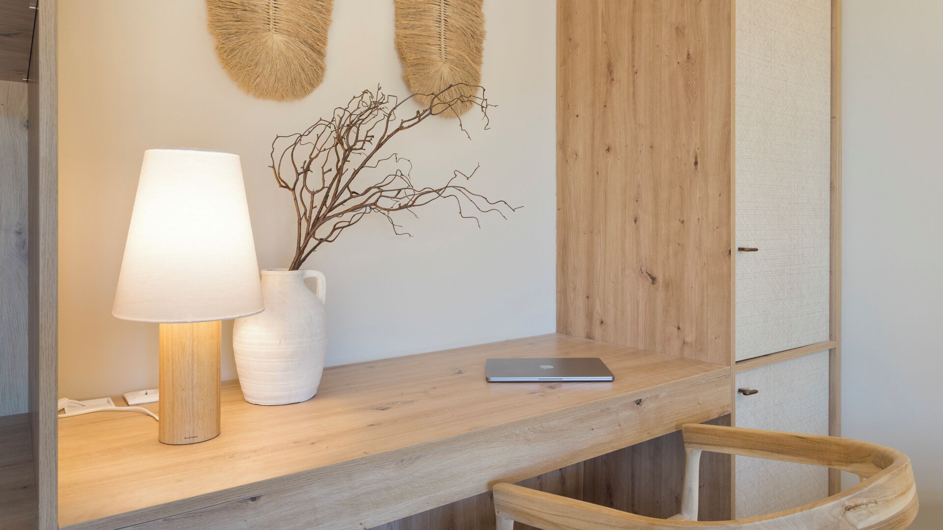 A wooden desk with a white lamp, a white vase with branches, and a laptop.