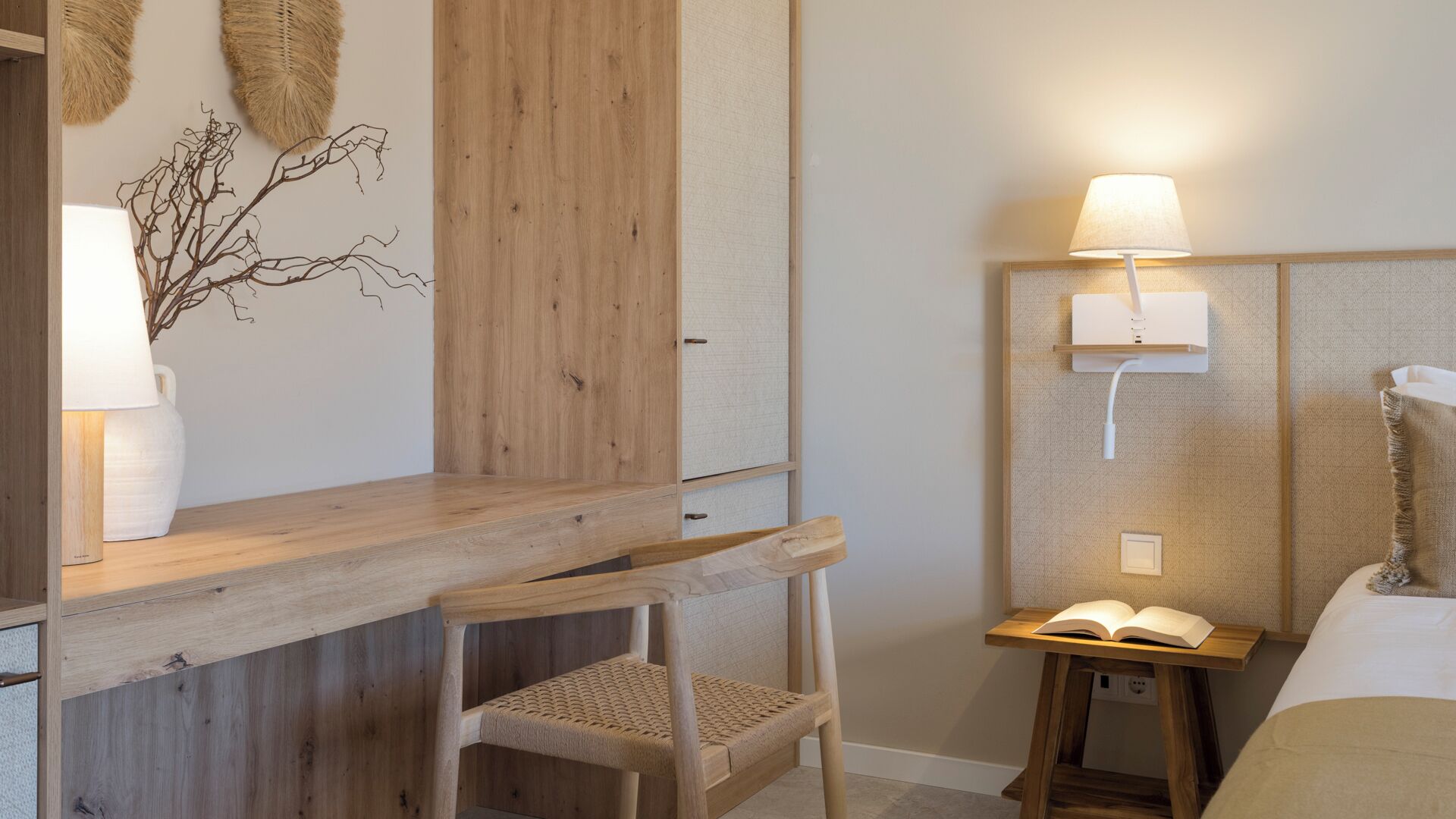 A room with a wooden desk, two chairs, a bedside table with a lamp, and a wall-mounted light fixture.