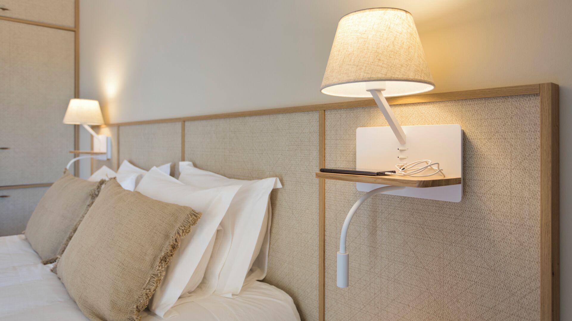 Une chambre d’hôtel avec un lit, deux lampes fixées au mur et une étagère sur laquelle se trouvent un stylo et des lunettes.