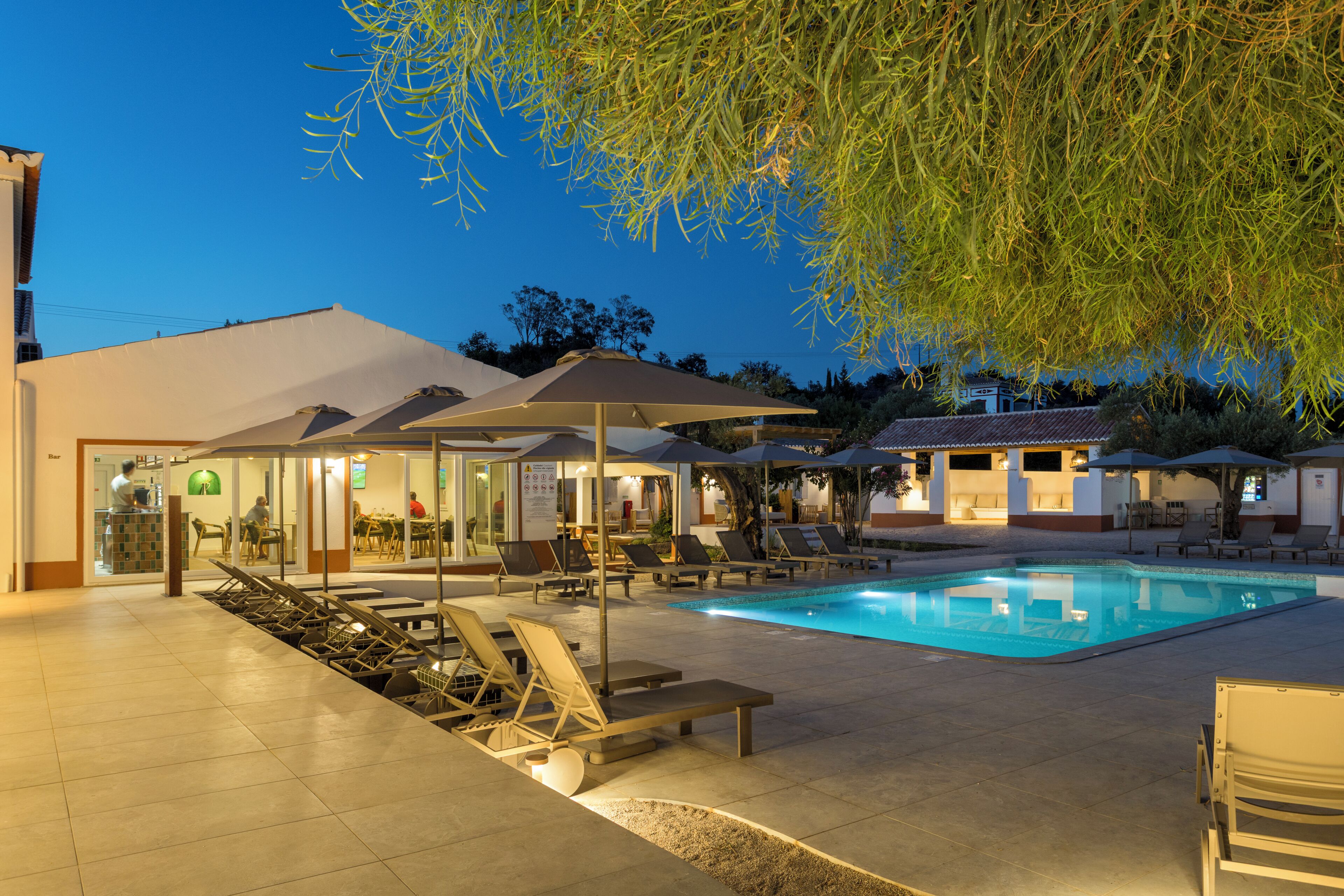 A swimming pool with lounge chairs and umbrellas, a bar area, and a building with a tiled roof.