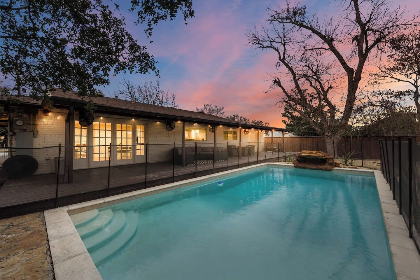 A swimming pool with a wooden deck and a fenced area, surrounded by trees and a building with windows and lights.