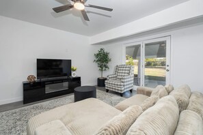 Spacious living room with hardwood floors, comfortable plush couch, a Smart TV with streaming services, and a ceiling fan. Enjoy fast Wi-Fi, natural light, and an open layout ideal for relaxing or remote work.