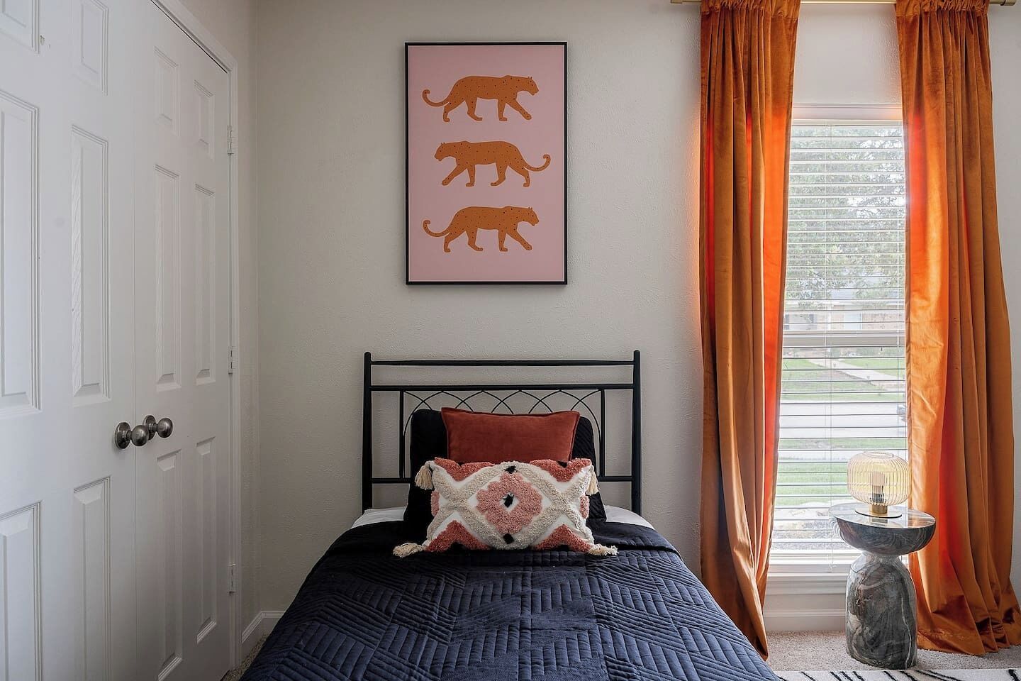 Une chambre à coucher avec un lit, un tableau représentant trois guépards accroché au mur, une fenêtre avec des rideaux et une table de chevet avec une lampe.