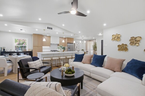 A modern living room with a sofa, coffee table, and chairs. There is a kitchen area with a refrigerator and cabinets.