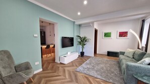 Ein modernes Wohnzimmer mit Fischgrätparkett, einem grauen Sofa, einem grauen Sessel, einem an der Wand montierten Flachbildfernseher, einer Topfpflanze und einem kleinen Essbereich im Hintergrund.