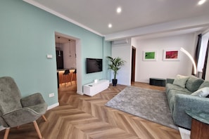 Ein modernes Wohnzimmer mit Fischgrätparkett, einem grauen Sofa, einem grauen Sessel, einem an der Wand montierten Flachbildfernseher, einer Topfpflanze und einem kleinen Essbereich im Hintergrund.