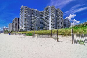 A beachfront high-rise building with multiple balconies, a sandy beach in the foreground, and a clear blue sky. - Oceanfront Condo with Sunset Views (Myrtle Beach)