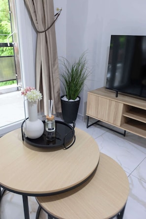 Ett rum med ett runt bord, en krukväxt, en vas med blommor, ett bricka med ett ljus och en tv-apparat som står på ett träställ.