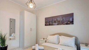 A hotel room with a bed, a cityscape painting, a wardrobe, a mirror, and a potted plant. - Fresh 1 Bd Apartment with Balcony in Thessaloniki (Thessaloniki)