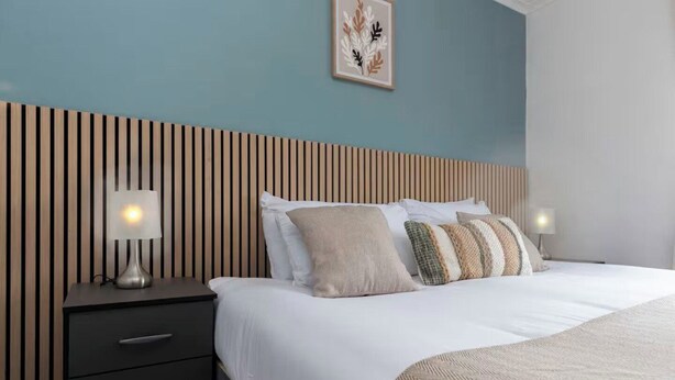 Una cama bien hecha con sábanas blancas y varios almohadones, una mesita de noche con lámpara y una pared con listones de madera verticales.