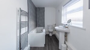 A modern bathroom with a bathtub, toilet, and sink, featuring a grey wall and a window with blinds. - *Discounts/Contractors/WIFI/Free Parking/Smart TV (Murton)