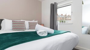 A neatly made bed with patterned pillows, a green throw blanket, and a window with a view of houses.