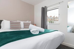 A neatly made bed with patterned pillows, a green throw blanket, and a window with a view of houses.