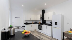 A modern kitchen with white cabinets, a black countertop, a stainless steel kettle, a microwave, a sink, and a bowl of fruit.