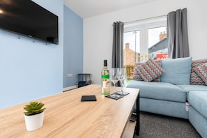 Ett modernt vardagsrum med en platt-TV som är monterad på väggen, ett träbord som används som soffbord med en växt, en vinflaska och två glas. Det finns en ljusblå soffa med mönstrade kuddar och en utsikt över omvärlden genom ett fönster med grå gardiner.