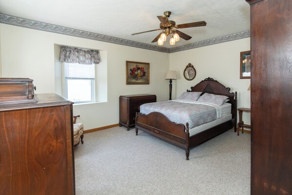 Une chambre à coucher avec un lit en bois, un ventilateur de plafond, une fenêtre avec un rideau, une table de chevet et une commode.