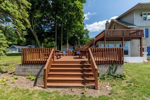 Un escalier en bois mène à une terrasse dotée de sièges extérieurs, le tout étant entouré d’arbres et d’un bâtiment aux fenêtres bleues.