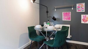Een eetruimte met een tafel voor vier personen, twee groene stoelen, een zwarte vloerlamp en bloemkunst in een lijst aan de muur.