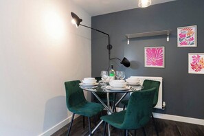 Ein Essbereich mit einem Tisch, gedeckt für vier Personen, zwei grüne Stühle, einer schwarzen Stehlampe und gerahmten Blumenbildern an der Wand.