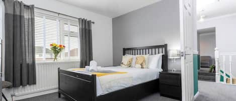 A hotel room with a black bed, white bedding, a bedside table with a lamp, a window with curtains, and a radiator.