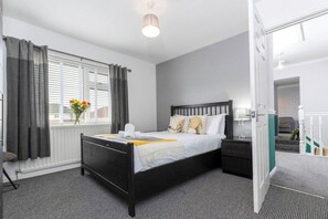 A hotel room with a black bed, white bedding, a bedside table with a lamp, a window with curtains, and a radiator.