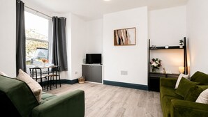 Een moderne woonkamer met een groene bank, een eethoek met een tafel en stoelen, een flatscreen televisie en een wandplank met decoratieve objecten.
