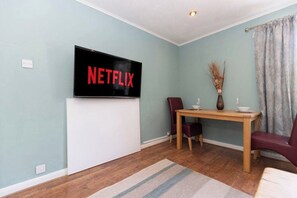 Ein Zimmer mit einem an der Wand befestigten Flachbildfernseher, einem Holzesstisch mit zwei Stühlen und einer Vase mit Trockenpflanzen darauf.