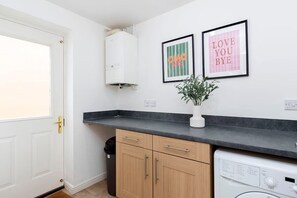 A kitchen with a white door, a black countertop, wooden cabinets, a washing machine, and two framed pictures on the wall. - Stylish Durham 3-Bed | Comfort & Long Stays (Spennymoor)