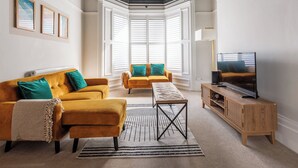A living room with a yellow sofa, a wooden coffee table, a TV stand, and a bay window with white shutters. - 4BR Contractor Stay | Parking, Wi-Fi, Spacious (Sunderland)