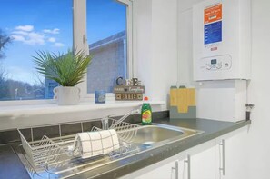 A kitchen with a dish rack, a potted plant, a water heater, and a window with a view of buildings. - Modern 2BR | Middlesbrough | Parking (Normanby)