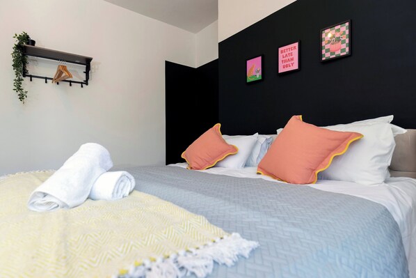 Una cama bien hecha con almohadas, una toalla y una manta. Hay una estantería con plantas y un marco de fotos en la pared.