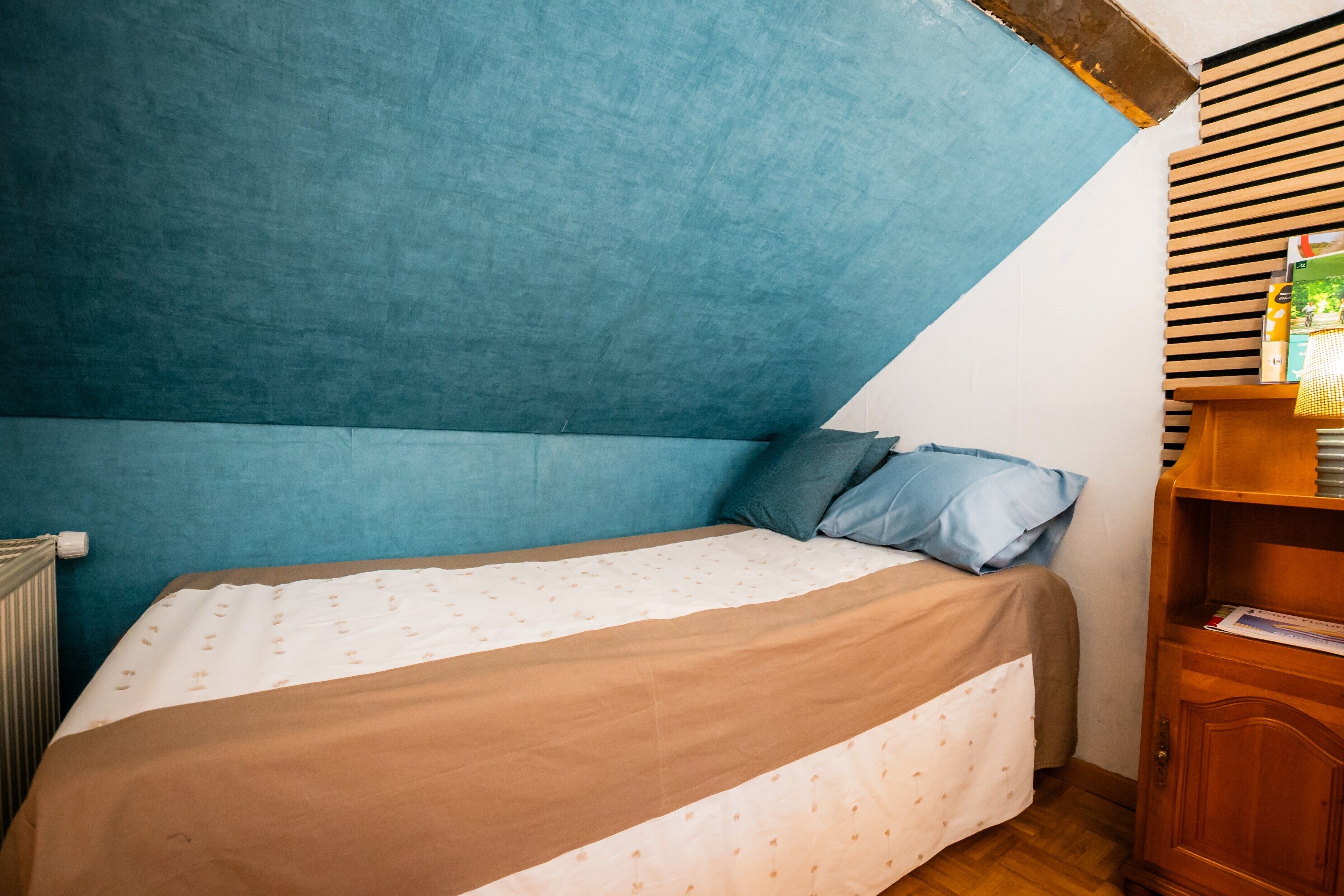 Quarto com teto inclinado, cama com colcha estampada, travesseiro azul e mesa de cabeceira de madeira com livros.
