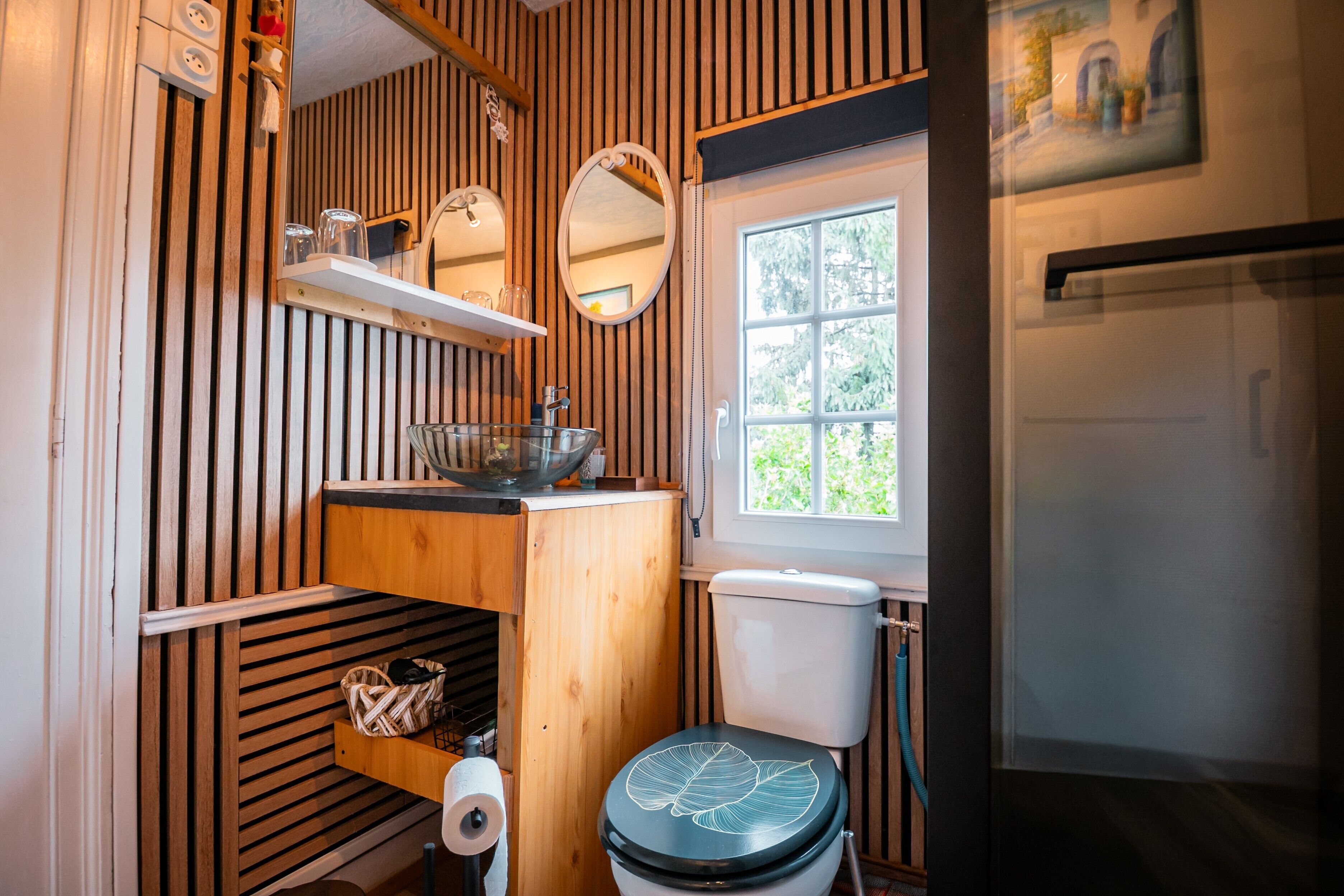 Een compacte badkamer met een wand van houten latten, een toilet met een bladmotief, een klein plankje met glazen en een raam met uitzicht.