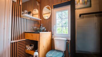 Een compacte badkamer met een wand van houten latten, een toilet met een bladmotief, een klein plankje met glazen en een raam met uitzicht.