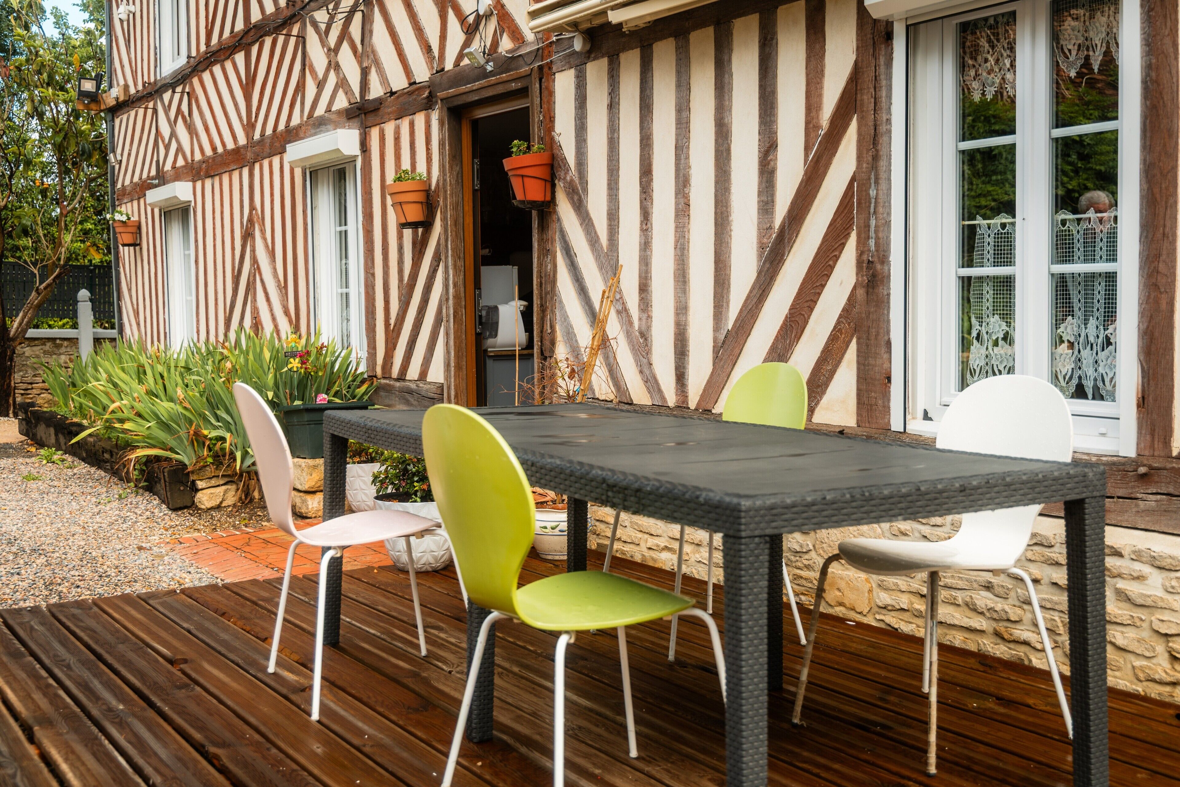 Une terrasse en bois avec une table et des chaises, un mur en pierre et une fenêtre ornée de rideaux en dentelle.