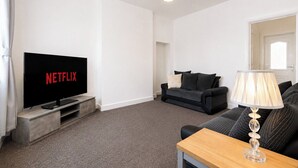 En stue med et fladskærms-tv der viser 'NETFLIX', en sofa, et sofabord og en lampe.