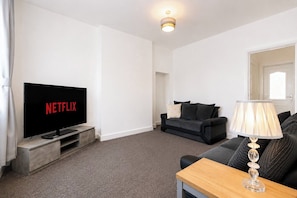 Ein Wohnzimmer mit einem Flachbildfernseher, der 'NETFLIX' anzeigt, einer Couch, einem Couchtisch und einer Lampe.