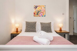 Ein Bett mit zwei gemusterten Kissen, einem gerollten weißen Handtuch, einer pinkfarbenen, gemusterten Bettdecke und einem farbenfrohen Kunstwerk darüber.