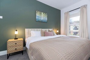 A bedroom with a bed, two bedside tables, a painting, and a window with curtains. - Durham 5BR | Contractors & Long Stay Discounts (Murton)