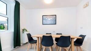 A room with a wooden table, black chairs, a framed picture on the wall, a window with curtains, and a plant on the windowsill. - Durham 5BR | Contractors & Long Stay Discounts (Murton)