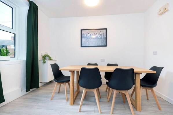A room with a wooden table, black chairs, a framed picture on the wall, a window with curtains, and a plant on the windowsill.