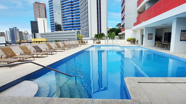 A rooftop pool with lounge chairs, a clear view of the city skyline, and a modern building with red and white exterior. - COCAR0116 - Boulevard Side Residencial (Salvador)
