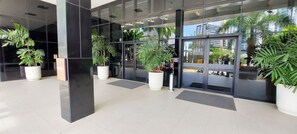 A modern building entrance with large glass doors, potted plants, and a tiled floor. - COCAR0116 - Boulevard Side Residencial (Salvador)