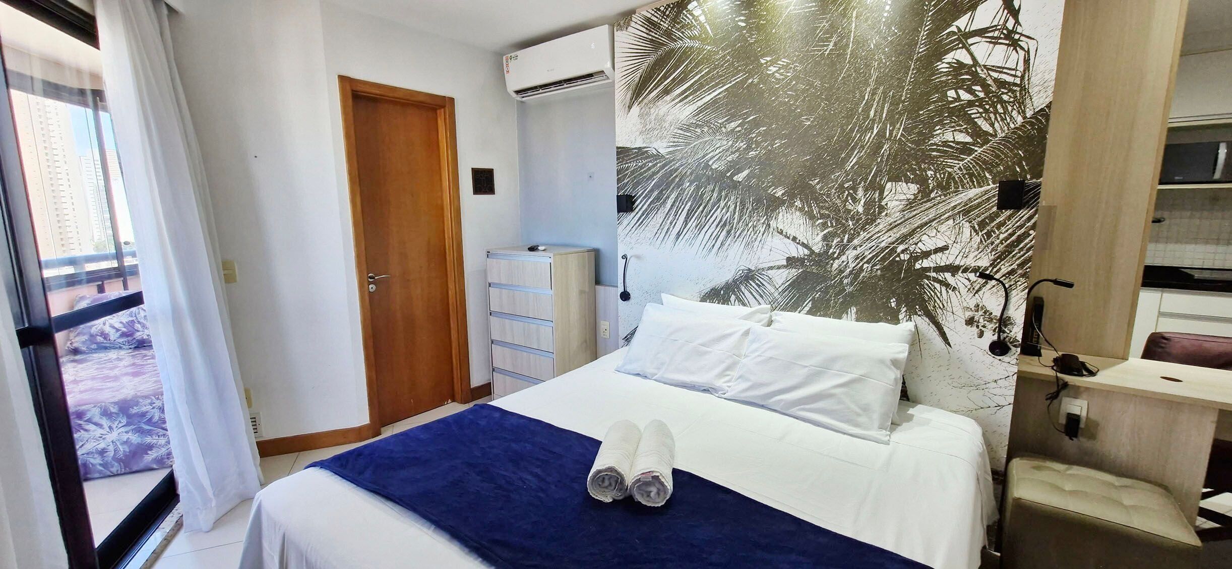 A hotel room with a bed, a wardrobe, an air conditioning unit, and a view through a window.